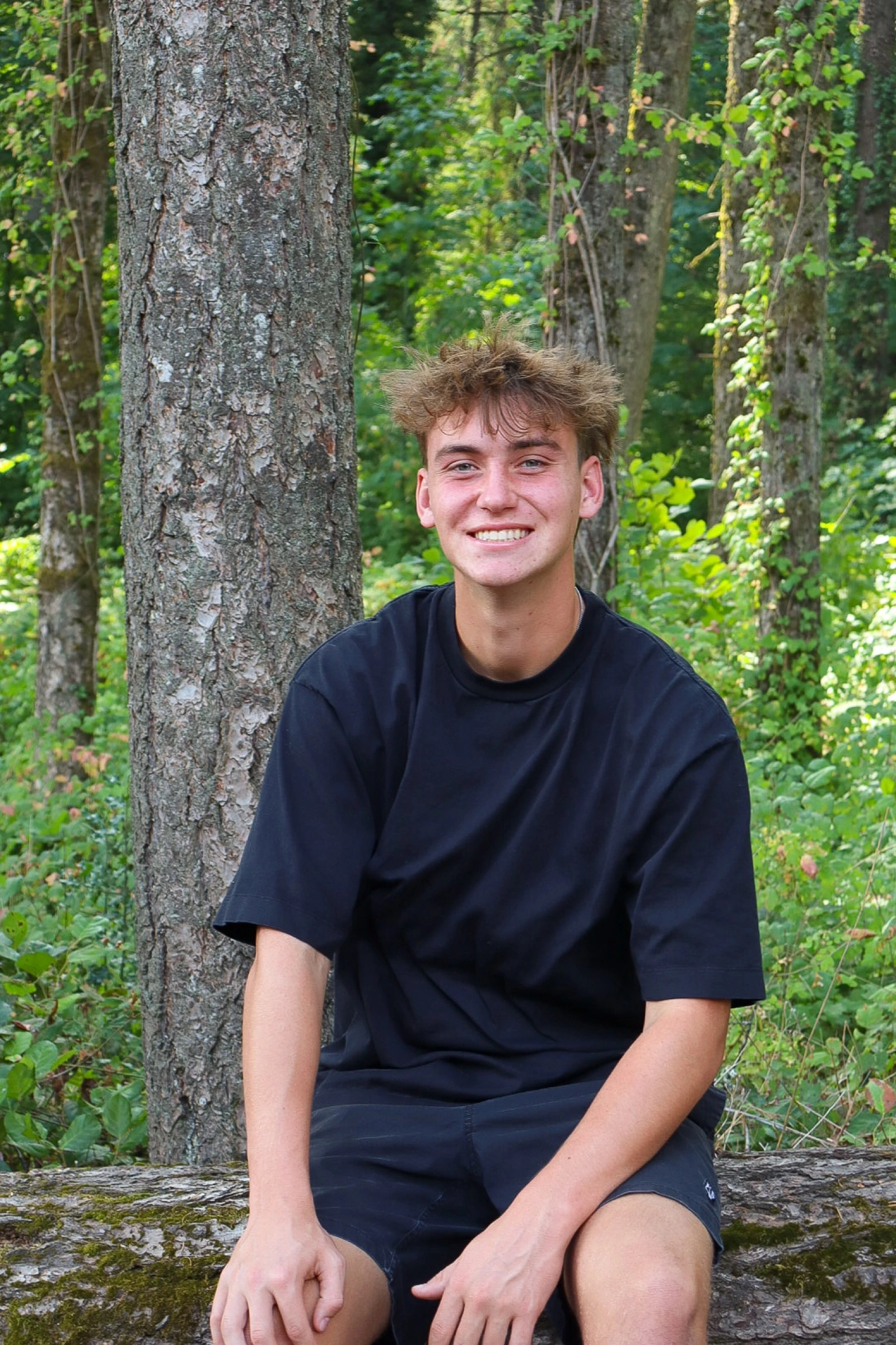 Young man smiling while seated on a mossy log in a forest